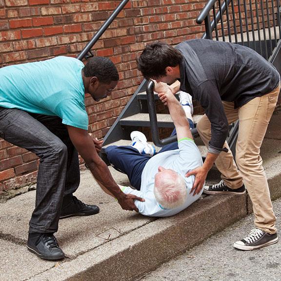 Elderly person that fell is on the floor and receiving assistance from two other men, who are helping him stand or walk.