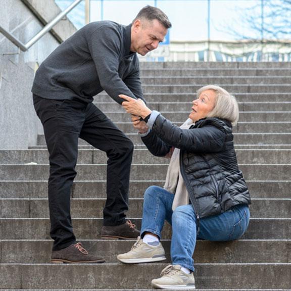 Man helping senior woman to get up while she is sitting on stairs outdoors in the city, holding her knee
