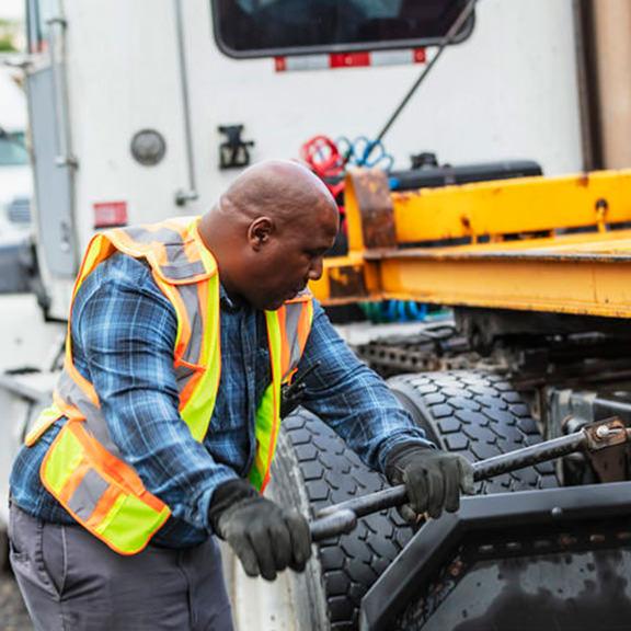 truck-accidents-02 A man in a safety vest is engaged in truck maintenance, inspecting the engine with tools in hand.